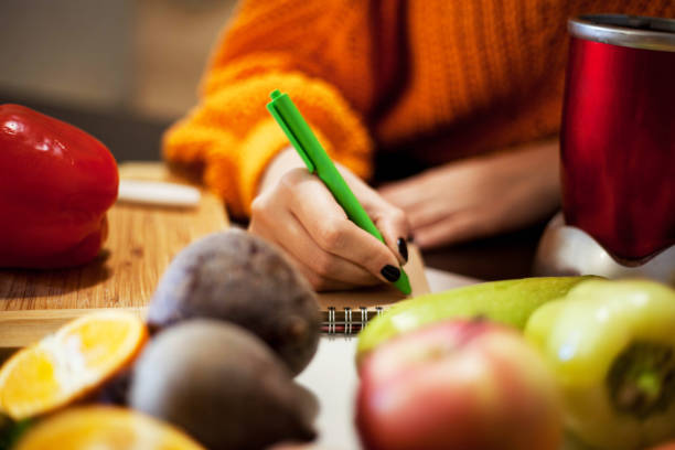 A notebook, pen and grocery list on a kitchen table beside a bowl of fruit